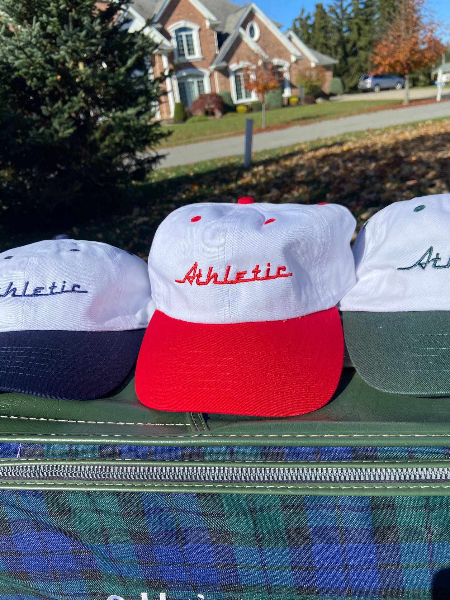Vintage style white hat with a red brim. Athletic and Affluent calls this colorway "Racing Red" and the AA club showcases their racing logo in this red color as well, reading "Athletic". It sits atop a vintage green leather luggage set on a fall day next to AA Club's Racer Hats in Tire Black and in Gas it Green