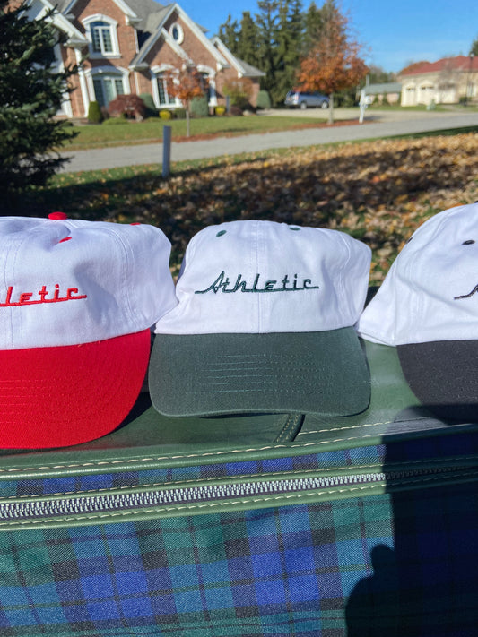 Vintage style white hat with a green brim. Athletic and Affluent calls this colorway "Gas It Green" and the AA club showcases their racing logo in gas it green font as well, reading "Athletic". It sits atop vintage plaid luggage between AA Clubs Racer Hats in "racing red" and "tire track black"