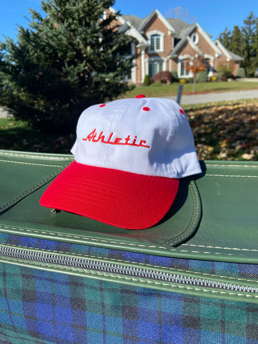 Vintage style white hat with a red brim. Athletic and Affluent calls this colorway "Racing Red" and the AA club showcases their racing logo in this red color as well, reading "Athletic". It sits atop a vintage green leather luggage set on a fall day
