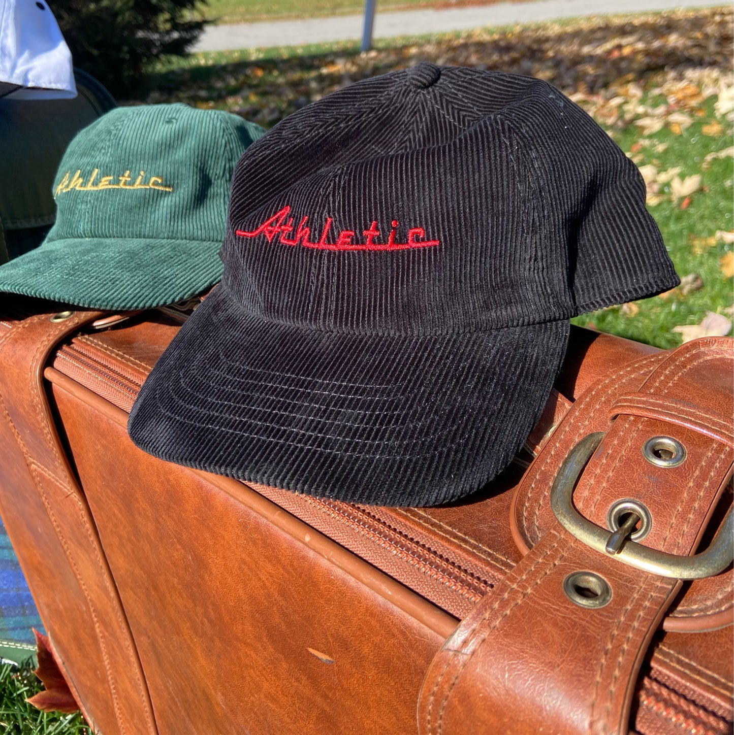 Black corduroy hat with red Athletic and Affluent branding spelling, "Athletic" in our classic racer font. It placed on a vintage leather suitcase on a bright fall morning next to AA Club's "Green Cord Cap"