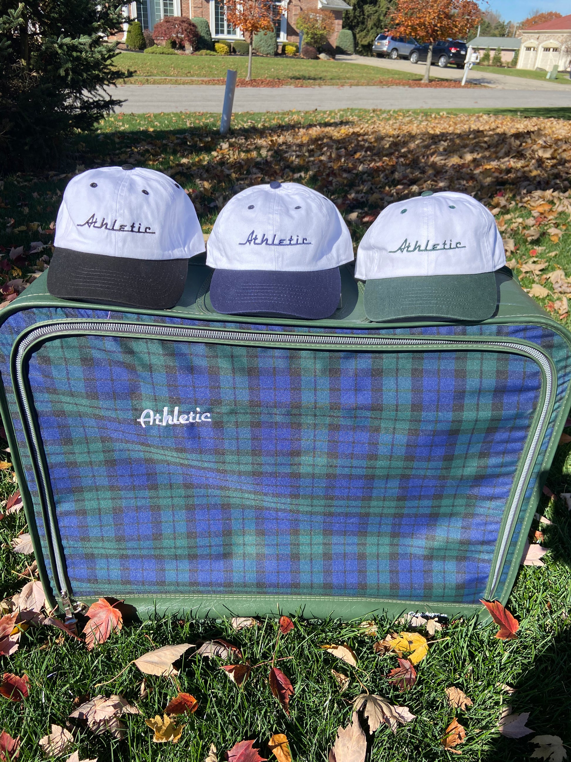 Vintage style white hat with a green brim. Athletic and Affluent calls this colorway "Gas It Green" and the AA club showcases their racing logo in gas it green font as well, reading "Athletic". It sits atop vintage plaid luggage between AA Clubs Racer Hats in "neutral navy" and "tire track black"