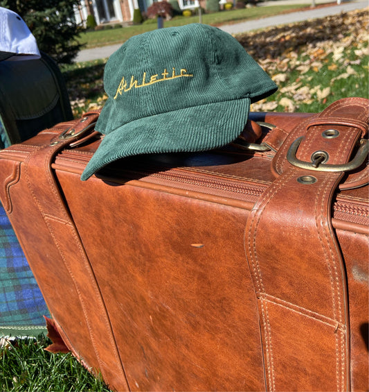 Green (forest green) corduroy hat with gold (yellow) Athletic and Affluent branding spelling, "Athletic" in our classic racer font. It placed on a vintage leather suitcase on a bright fall morning.