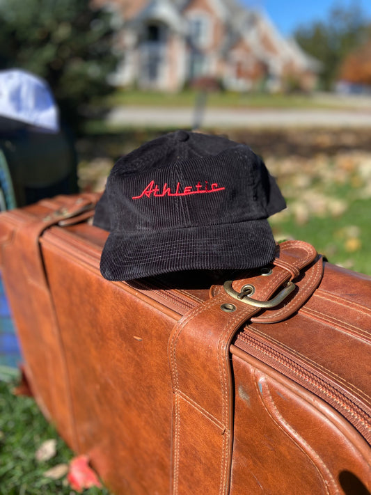 Black corduroy hat with red Athletic and Affluent branding spelling, "Athletic" in our classic racer font. It placed on a vintage leather suitcase on a bright fall morning. 
