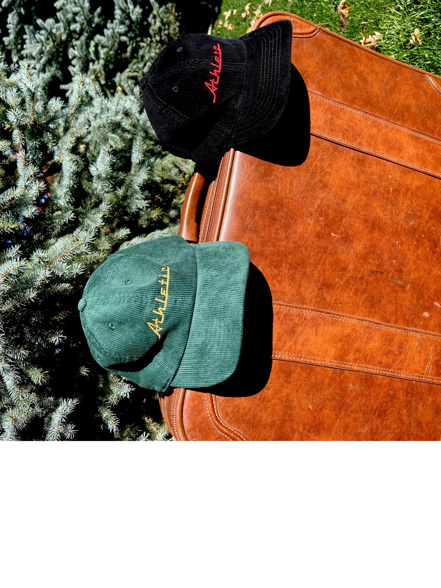 Green (forest green) corduroy hat with gold (yellow) Athletic and Affluent branding spelling, "Athletic" in our classic racer font. It placed on a vintage leather suitcase on a bright fall morning next to AA Club's "Black Cord Cap"