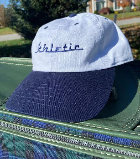 Vintage style white hat with a navy brim. Athletic and Affluent calls this colorway "racer Navy" and the AA club showcases their racing logo in a navy as well, reading "Athletic". It sits atop a vintage green leather luggage set on a fall day