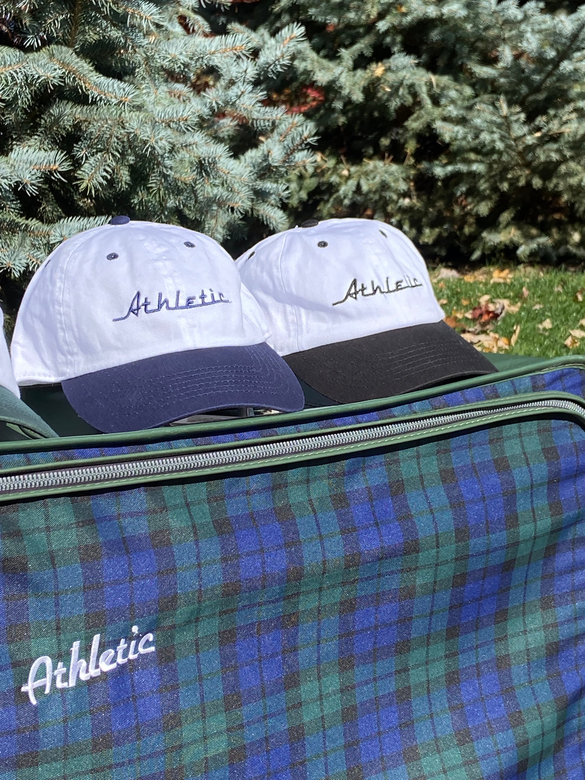 Vintage style white hat with a navy brim. Athletic and Affluent calls this colorway "racer Navy" and the AA club showcases their racing logo in a navy as well, reading "Athletic". It sits atop a plaid logo luggage set next to AA Clubs' Racer Hat in Tire Black