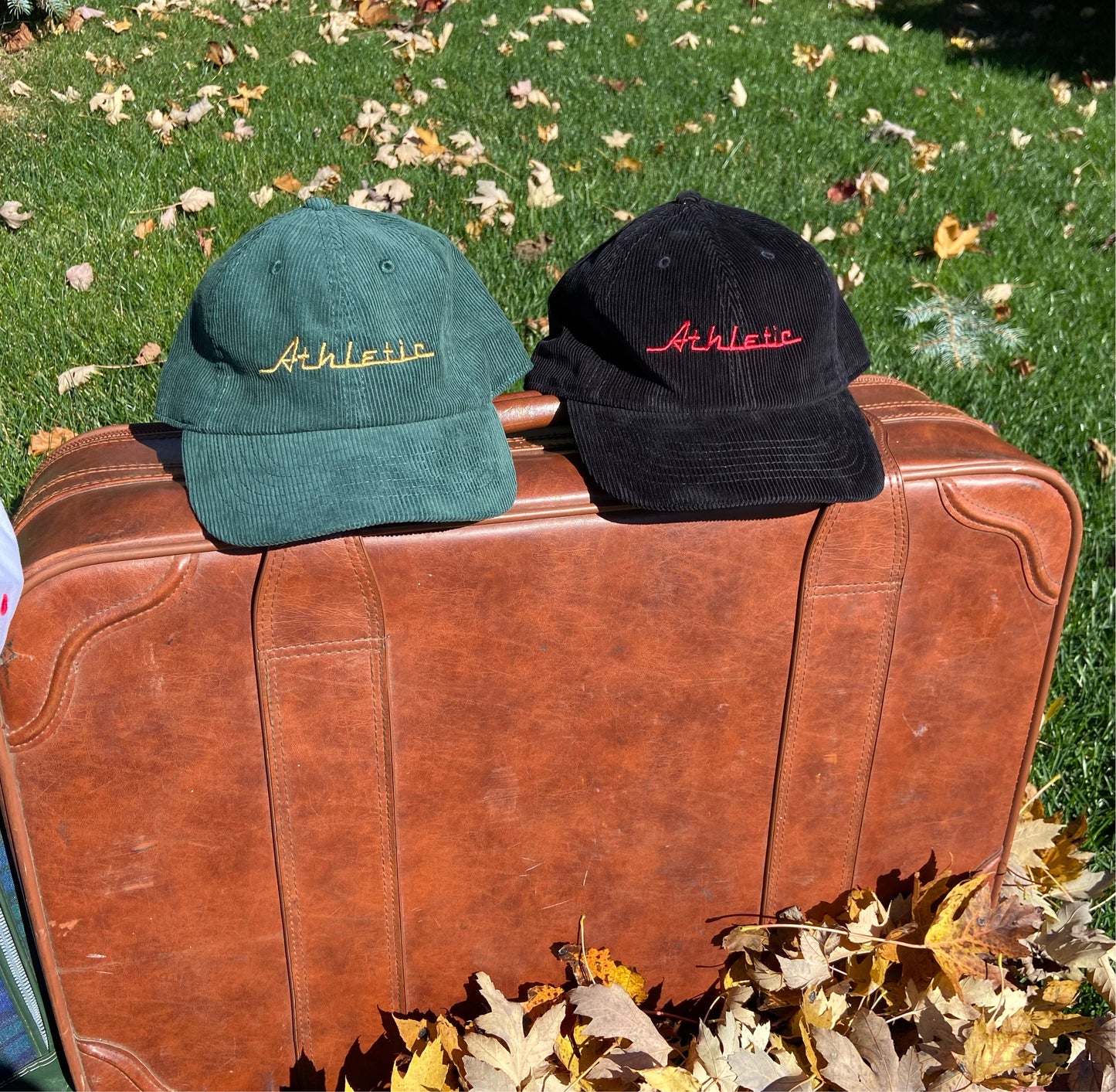 Green (forest green) corduroy hat with gold (yellow) Athletic and Affluent branding spelling, "Athletic" in our classic racer font. It placed on a vintage leather suitcase on a bright fall morning next to AA Club's "Black Cord Cap"