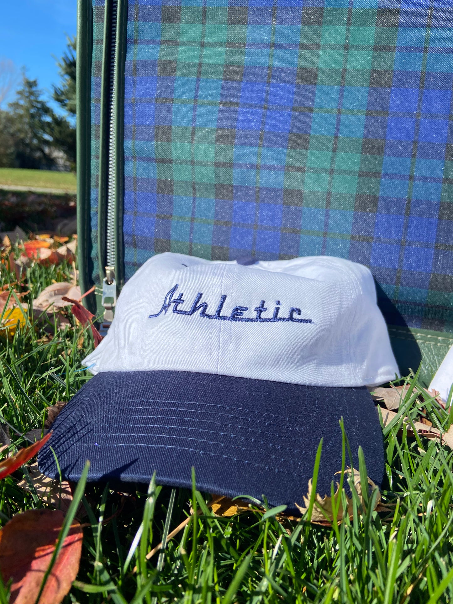 Vintage style white hat with a navy brim. Athletic and Affluent calls this colorway "Navy" and the AA club showcases their racing logo in a navy as well, reading "Athletic". It sits near a vintage plaid suitcase in the lush fall grass.