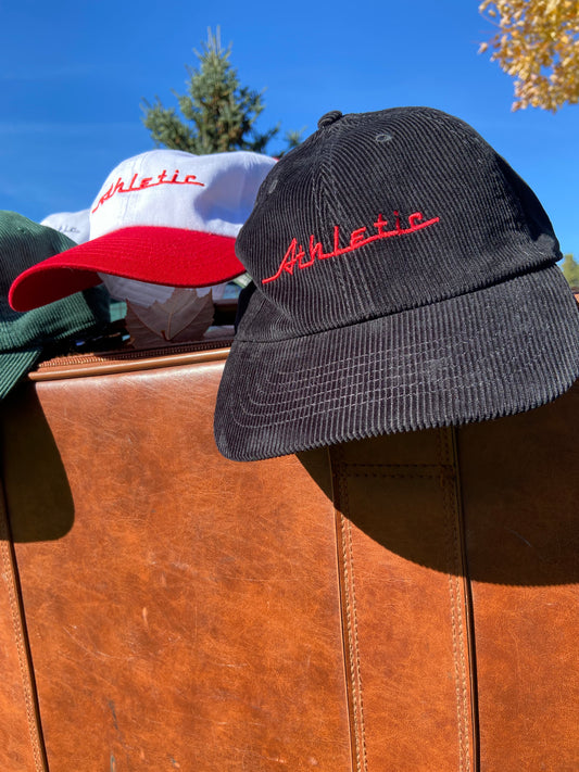 Black corduroy hat with red Athletic and Affluent branding spelling, "Athletic" in our classic racer font. It placed on a vintage leather suitcase on a bright fall morning next to AA Club's Racer hat in "Racing Red"