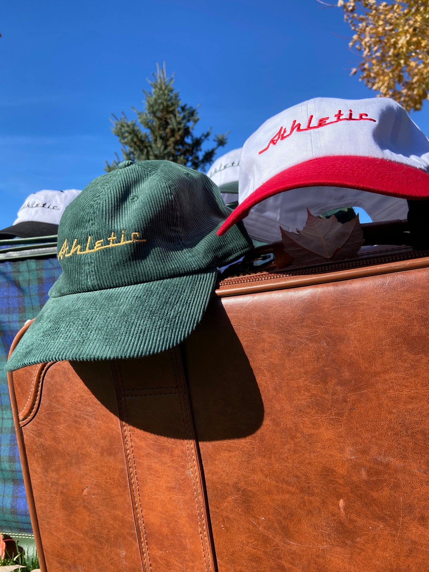 Green (forest green) corduroy hat with gold (yellow) Athletic and Affluent branding spelling, "Athletic" in our classic racer font. It placed on a vintage leather suitcase on a bright fall morning next to AA Club's Racer Hat in "Racing Red"