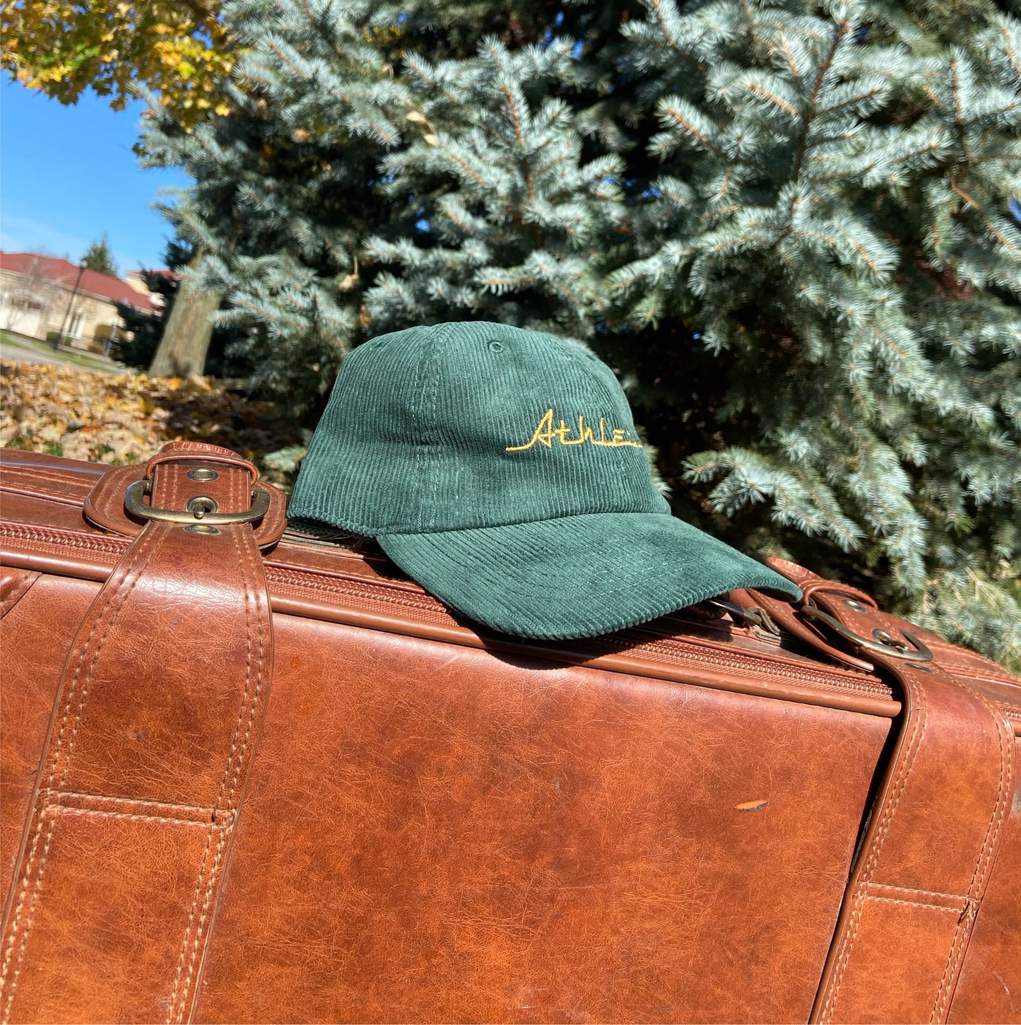 Green (forest green) corduroy hat with gold (yellow) Athletic and Affluent branding spelling, "Athletic" in our classic racer font. It placed on a vintage leather suitcase on a bright fall morning.