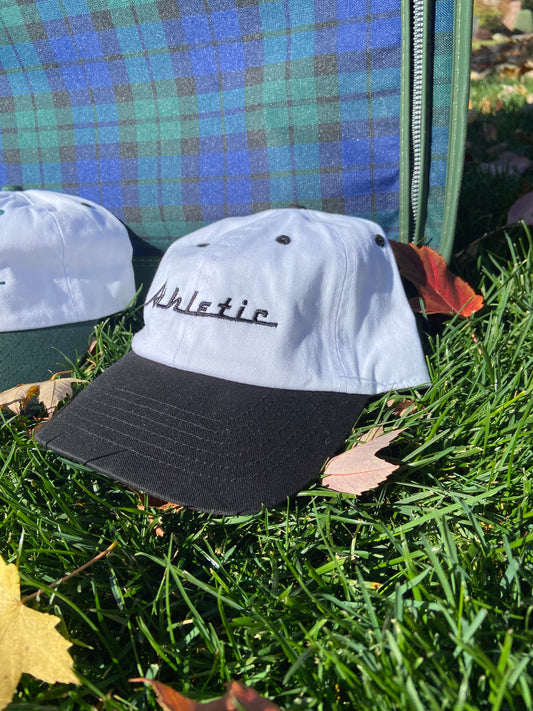 Vintage style white hat with a black brim. Athletic and Affluent calls this colorway "tire black" and the AA club showcases their racing logo in a tire black font as well, reading "Athletic". It sits in front of a vintage plaid luggage.