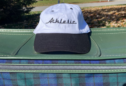 Vintage style white hat with a black brim. Athletic and Affluent calls this colorway "tire black" and the AA club showcases their racing logo in a tire track black font as well, reading "Athletic". It sits atop a vintage leather plaid luggage set in front of a pile of fall leaves.