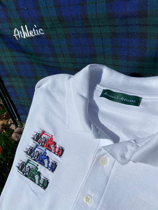 Casual classy vintage style white polo with Athletic and Affluent Race Team embroidery. Red blue and green racecars are displayed on the crisp white polo in front of an old-school plaid suitcase and some fall leaves. 