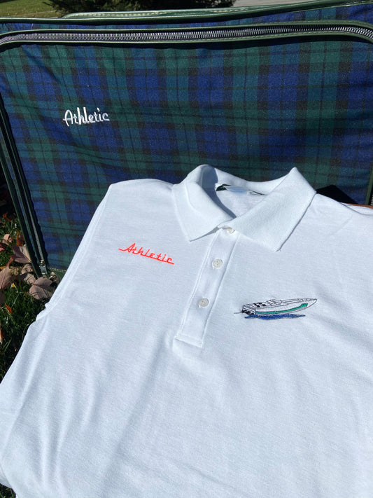 Casual classy white polo with a vintage style green-striped racing boat. The AA Club logo is displayed in pink spelling "Athletic" and this vintage polo is placed in front of a preppy plaid suitcase on a sunny fall day.