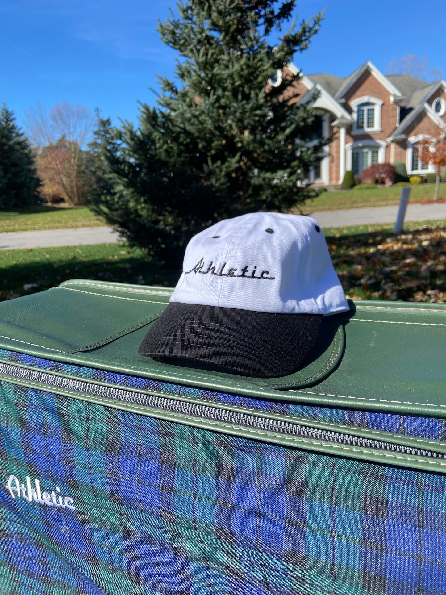 Vintage style white hat with a black brim. Athletic and Affluent calls this colorway "tire black" and the AA club showcases their racing logo in a tire black font as well, reading "Athletic". It sits atop an AA Club embroidered vintage leather plaid luggage set in front of a pile of fall leaves.